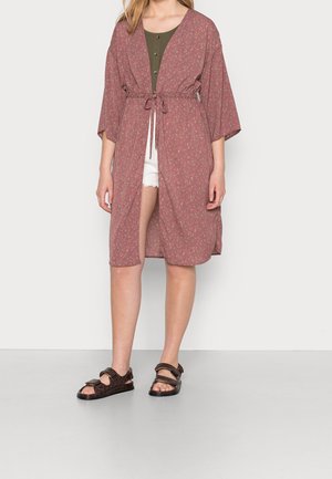 Mujer con bata floreada rosa hasta la rodilla con cinturón en la cintura, top verde oliva, pantalones cortos blancos y sandalias marrón oscuro, de pie sobre un fondo liso.