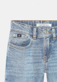 Jasnoniebieskie dżinsy z przodu z kieszeniami, pomarańczowym przeszyciem, srebrnym guzikiem z wygrawerowanym napisem "cK" oraz metką Calvin Klein Jeans wszytą w pasek.