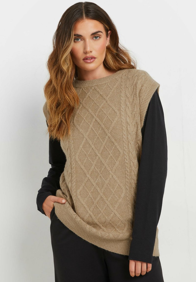 Maglione senza maniche in cotone beige con un motivo a trecce, indossato sopra una maglietta nera a maniche lunghe. Il maglione ha un collo rotondo e bordi a coste.
