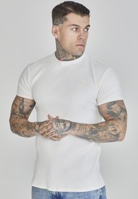 Tricou alb cu dungi, cu mâneci scurte, având un guler rotund și material texturat. Purtați-l peste blugi albaștri, cu tatuaje detaliate vizibile pe brațe.