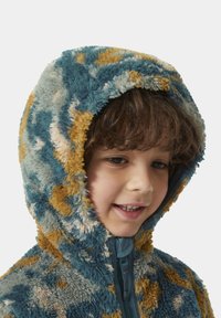 Giacca a cappuccio morbida e multicolore con tonalità di blu, giallo e beige; texture soffice con chiusura a zip sul davanti.