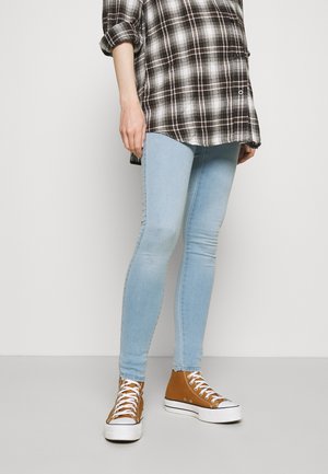 Persona con jeans ajustados azul claro, zapatillas altas marrones y una camisa de cuadros en blanco y negro con las mangas remangadas.