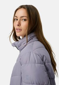 Lichtpaarse puffervest met een hoge kraag, gewatteerde textuur en een ritssluiting. Bevat zichtbare stiksels en een logo patch op de mouw.