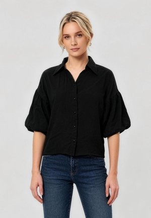 Kvinde med blond hår iført en sort skjorte med knapper og puffede halvlange ærmer og blå jeans, stående mod en lysegrå baggrund.