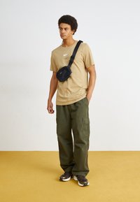 Beige T-krekls ar baltu logo, melna plecu soma, zaļi kravas bikses un tumši treneri. Modelis stāv uz dzeltena grīdas pret gaismu sienu.