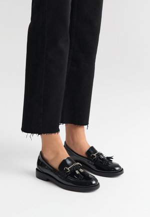 Black patent leather loafers with tassels and metal hardware accents, paired with cropped black trousers featuring frayed hem.