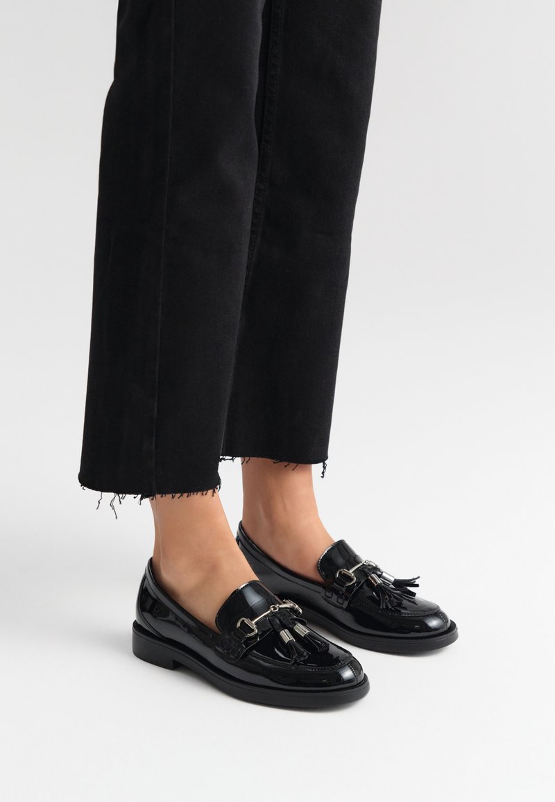Mustad nahast loafers, millel on pomponid ja metallist aksessuaarid, koos lühikeste mustade pükstega, millel on erkääred.