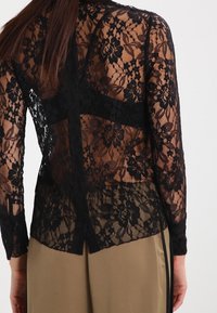Top floreale in pizzo nero a maniche lunghe indossato sopra un sottocutaneo nero, abbinato a pantaloni beige, visto da dietro.