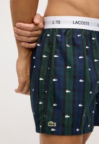 Kostkované boxerky Lacoste v zelené, námořnické a černé barvě, zdobené bílými vzory krokodýla a bílým elastickým pasem s textem "LACOSTE".