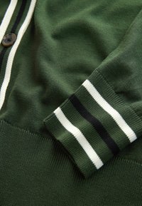 Zeleni pleteni kardigan s kopčanjem na gumbe, s rebrastim manšetama u bijelim i crnim prugama, te glatkom, mekanom teksturom.