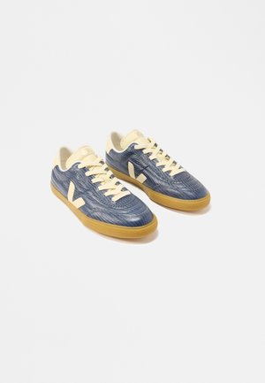 Zapatillas bajas texturizadas en azul con cordones y detalles en crema, suelas de goma color caramelo, mostradas lado a lado sobre un fondo blanco.