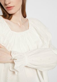 Blusa de algodão branca com decote franzido e mangas longas, apresentando detalhes em broderie anglaise e um ajuste descontraído. Superfície texturada.