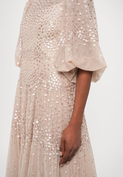 Sukienka z cekinami w jasnym beżu z teksturowanym, przezroczystym materiałem. Posiada bufiaste rękawy oraz ozdoby w różnych rozmiarach na korpusie.