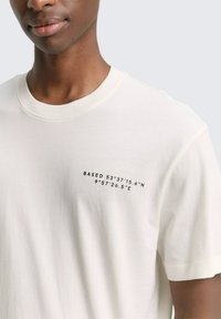 Licht beige katoenen t-shirt met korte mouwen, voorzien van een zwarte afgedrukte tekst aan de linkerzijde die luidt: "BASED 53°37'15.6"N 9°57'26.5"E."