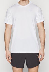 Vit atletisk t-shirt med en subtil textur, rund hals, korta ärmar och en ljusblå logotyp på bröstet. Matchas med mörka shorts.