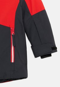 Rukáv černé a červené zimní bundy dítěte s červeným zipem a nastavitelným suchým zipem na bílém pozadí.