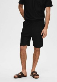 Juodi ripsuoti šortai su elastinga juosmeniu, derinami su atitinkančiu trumpų rankovių viršumi ir ruda šlepetėmis. Atsitiktinė ir patogi išvaizda.