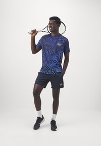 Conjunto deportivo para hombres que incluye una camiseta de manga corta con patrón de remolinos azules y pantalones cortos en color marino, combinados con zapatillas negras y calcetines blancos.