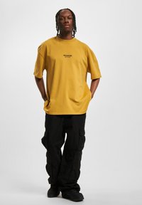 Gul bomull T-skjorte med rund hals og korte ermer, med svart tekst "PEGADOR" på brystet. Kombinert med svarte cargo-bukser.