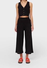 Kvinna i svart ärmlös knäppt crop-top, vida cropped byxor och ljusorange sandaler mot en neutral bakgrund.