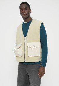 Gilet in pile beige chiaro, con due tasche frontali dotate di patte in tessuto beige. Il design presenta una scollatura a V profonda e una cerniera centrale.