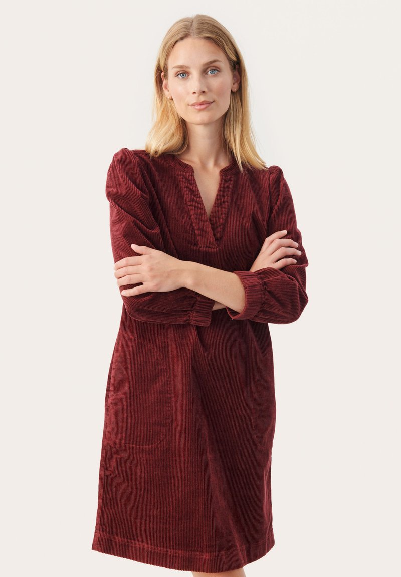 Rochie din catifea cu un ton roșu închis, având decolteu în V, mâneci lungi cu volum și buzunare laterale. Textura este striată și moale, având o croială lejeră.