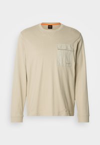 Béžová košeľa s dlhým rukávom z mäkkého materiálu, s textúrovanou prednou vreckom, okrúhlym výstrihom a jemnou značkou na hrudi.