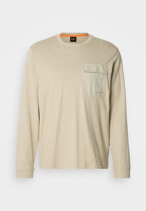 Beżowa koszula z długim rękawem wykonana z miękkiego materiału, z teksturowaną przednią kieszenią, okrągłym dekoltem i subtelnym logo na piersi.