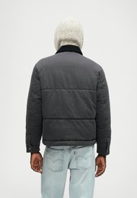 Jachetă puffer din țesătură gri, cu un design matlasat, guler negru și o croială lejeră, asortată cu blugi de un albastru deschis.