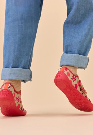 Kind trägt hochgekrempelte blaue Jeans, beiger Socken und rote Schuhe mit Gummisohle, verziert mit Eulen- und Herzmustern, steht auf beigem Boden.