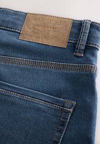 Primo piano della tasca posteriore di jeans in denim blu con cuciture arancioni e una patch rettangolare in pelle marrone con la scritta "Denim Supply."