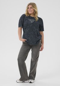 Black marble-patterned t-shirt with short sleeves, curved hem, featuring a white logo. Paired with gray, straight-leg jeans and white sneakers.