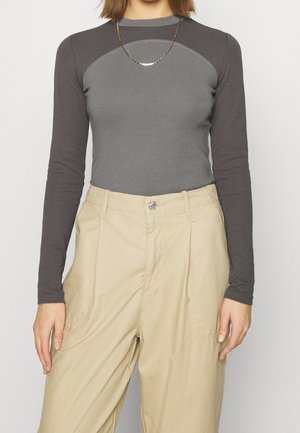 Kvinde iført en tætsiddende tofarvet grå langærmet skjorte, beige højtaljede bukser og en tynd guldkæde, stående foran en ensfarvet baggrund.