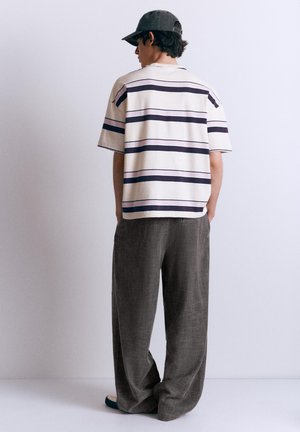 Young man wearing a white short-sleeve shirt with black stripes, loose dark gray pants, gray cap, standing with hands in pockets facing away.