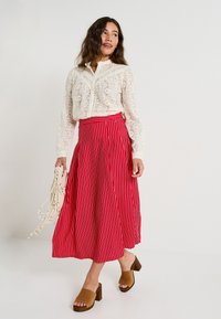 Blusa de encaje en off-white, con intrincados patrones florales; falda roja con rayas blancas verticales; sandalias de tacón cuadrado marrones; bolso crema.