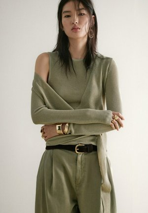 Donna che indossa un top a strati in maglia verde oliva con spalla tagliata, pantaloni abbinati, cintura nera e gioielli dorati su sfondo uniforme.