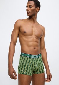 Πράσινα μπόξερ με γεωμετρικό σχέδιο, ελαστική μέση με το λογότυπο "LACOSTE" σε ναυτικό μπλε και λευκές αποχρώσεις.