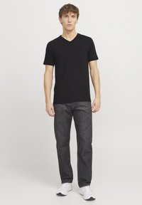 T-shirt noir à col en V associé à un jean droit de lavage foncé. Le modèle porte des chaussures de sport blanches, se tenant devant un fond uni.