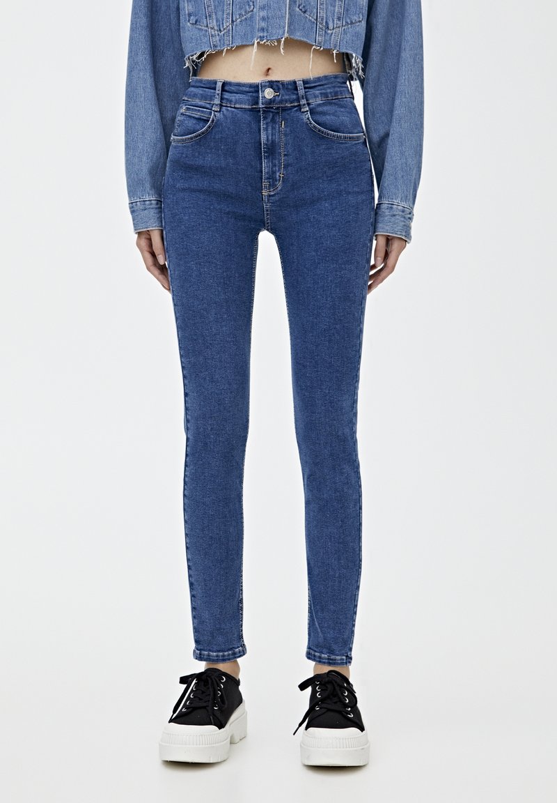 Jeans ajustados de mezclilla azul con cintura alta, corte slim y diseño de cinco bolsillos. Combinados con zapatillas negras con suelas blancas.