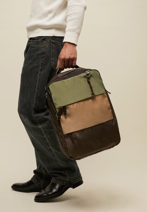 Persoon draagt zwarte jeans, witte trui en zwarte schoenen en houdt een rechthoekige driekleurige tas vast bij het bovenste handvat.