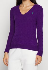 Pull en maille torsadée violet avec un décolleté en V et des poignets côtelés. Présente un petit logo brodé sur la poitrine. Texture douce.