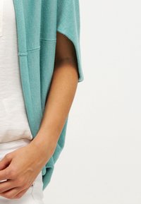 Lätt, teal cardigan med öppen front, korta ärmar och en mjuk textur, som bärs tillsammans med en vit topp och ljusa byxor.