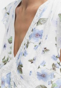 Blumenkleid aus weißem, durchsichtigem Stoff mit blauen Blumenmustern. Spitzendetails entlang des V-Ausschnitts und der Ärmel sorgen für Textur und Detail.