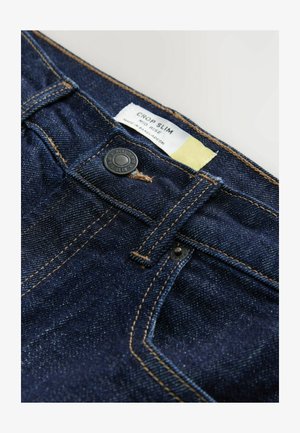 Jeans in denim scuro con un design crop slim, cuciture in rame e un bottone nero. L'etichetta indica "CROP SLIM, MID RISE" prodotto in Bangladesh.