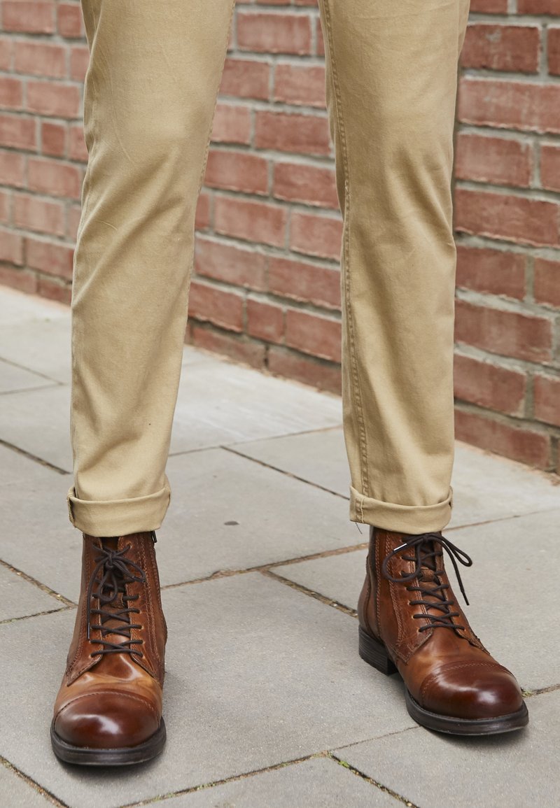 Botas de tobillo de cuero marrón con punta redonda, paneles laterales texturizados y diseño de cordones, combinadas con pantalones caqui enrollados sobre una superficie de ladrillos.