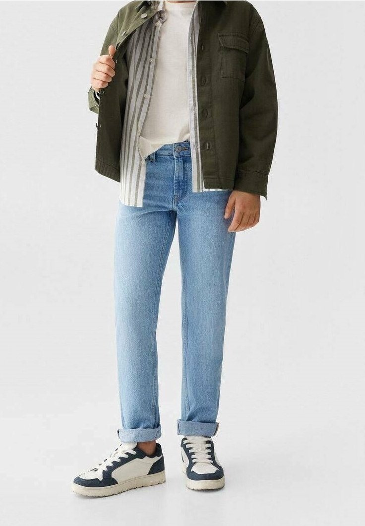 Personne portant un jean bleu clair retroussé, des baskets blanches avec des touches sombres, une chemise blanche, une chemise rayée à boutons et une veste vert foncé.
