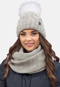 Bonnet en maille gris clair avec un pompon en fourrure blanche sur le dessus, associé à une écharpe infini en tricot gris clair épaisse. Matériaux texturés.