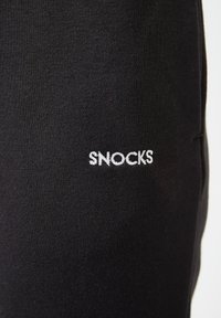 Zwarte katoenen stof met een gladde textuur met een witte geborduurde "SNOCKS" logo. Close-up van het been gedeelte van het kledingstuk.