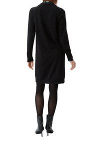 Schwarzes gestricktes Kleid mit langen Ärmeln und geripptem Kragen. Getragen mit schwarzen, knielangen Stilettos und transparenten schwarzen Strumpfhosen.