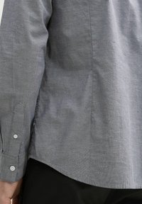 Camicia grigia con bottoni in tessuto testurizzato, caratterizzata da un colletto a punta, maniche lunghe e orlo curvo. I bottoni sono di colore chiaro.
